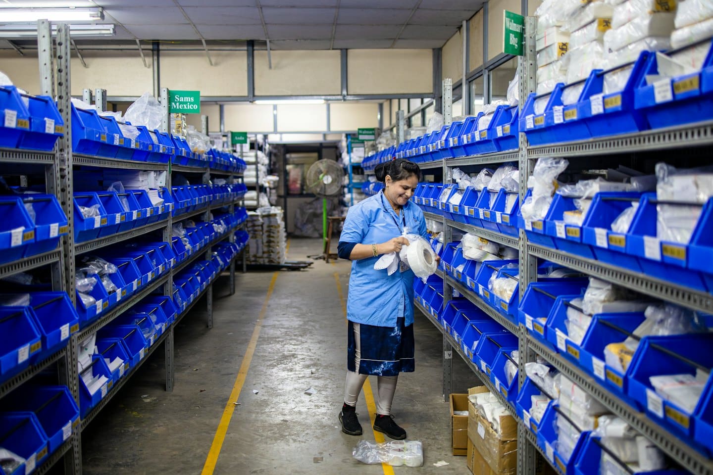 Kobieta w niebieskim uniformie sortuje przedmioty z plastikowych pojemników w magazynie.
